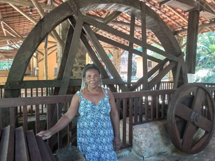 A líder quilombola Laura de Jesus Braga está em frente ao máquinário, de mais de um século de idade, responsavel pela produção de farinha que era a principal fonte de renda do Quilombo da Fazenda, local da fotografia.