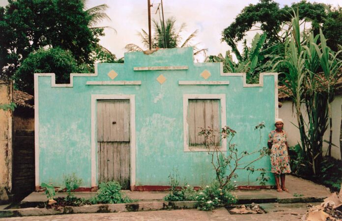 Representando a arquitetura sertaneja exibida em documentário lançado no Festival Internacional, a imagem mostra uma casa no sertão, com fachada azul, uma porte e uma janela. Junto a ela, uma mulher, de vestido e com lenço na cabeça.