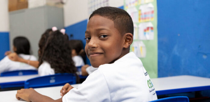 Criança negra sorrindo em frente à câmera. Atrás dele, há alunos virados para o outro lado. Todos estão em um ambiente escolar.