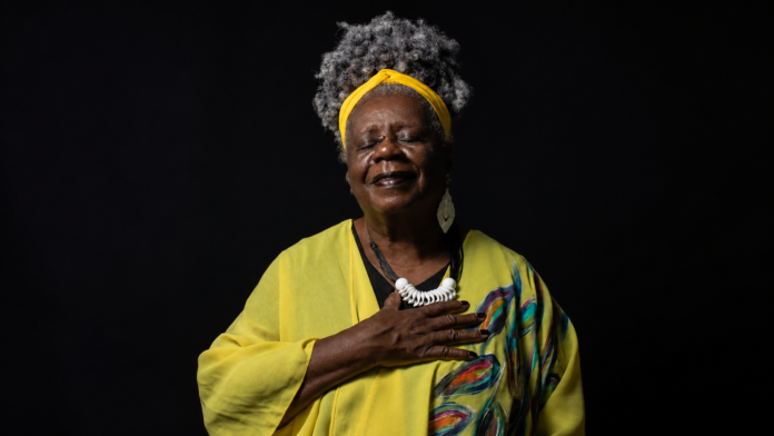 Retrato horizontal de uma mulher negra idosa, de olhos fechados e mão sobre o peito, usando roupa amarela, faixa na cabeça e colar branco, diante de fundo preto