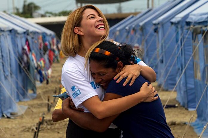 Alt text: Mulher com camiseta da ONU abraça outra mulher em uma área com tendas de apoio humanitário. Descrição: Voluntária da ONU abraça uma mulher em uma área de atendimento humanitário, com tendas ao fundo. Crédito: VNU Palavras-chave: voluntariado, ONU, ação humanitária, solidariedade, ajuda humanitária, trabalho voluntário, apoio social, Dia do Voluntariado