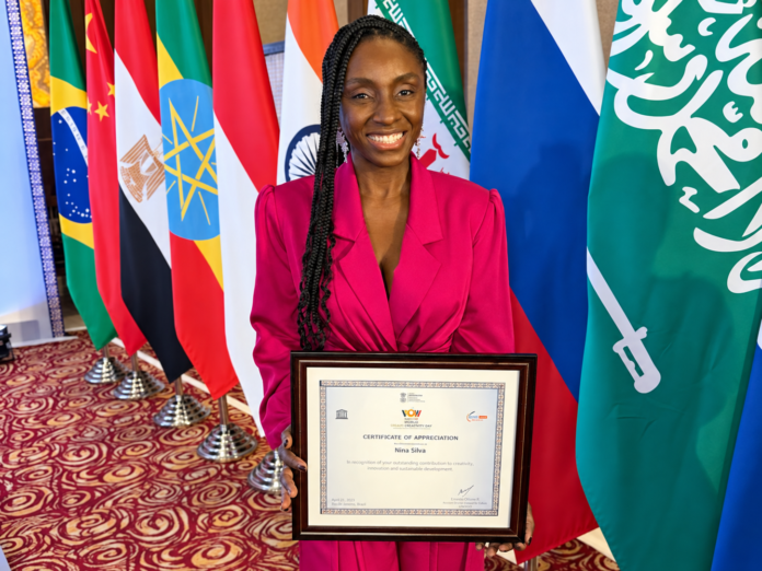 Nina Silva sorri para a câmera segurando um quadro, diante de bandeiras de países do BRICS e parceiros, em evento realizado em Nova Délhi, na Índia